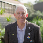 A man standing in a garden. His jacket is adorned with LGBTQIA+ pins and volunteering pins.