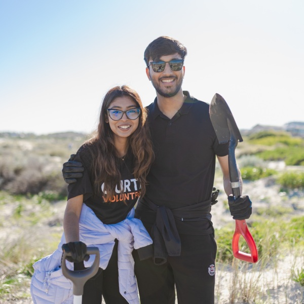 Two student volunteers holding shovels, on the sand dunes of a beach.