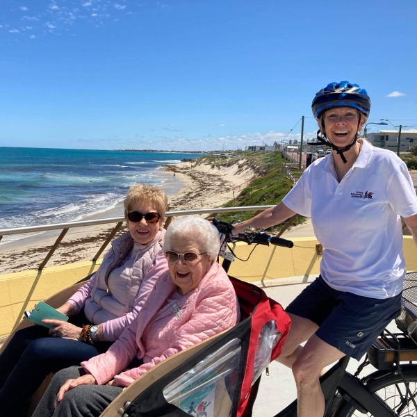Volunteer with helmet pedalling on bike and pushing two old women who are seated on a chair with wheels