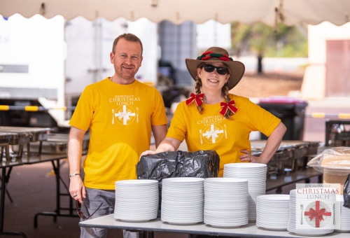 Two volunteers stand behind stacks of plates for a community lunch