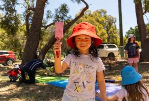 A young volunteer holding up a paintbrush. People are painting the footpath in the background behind her.